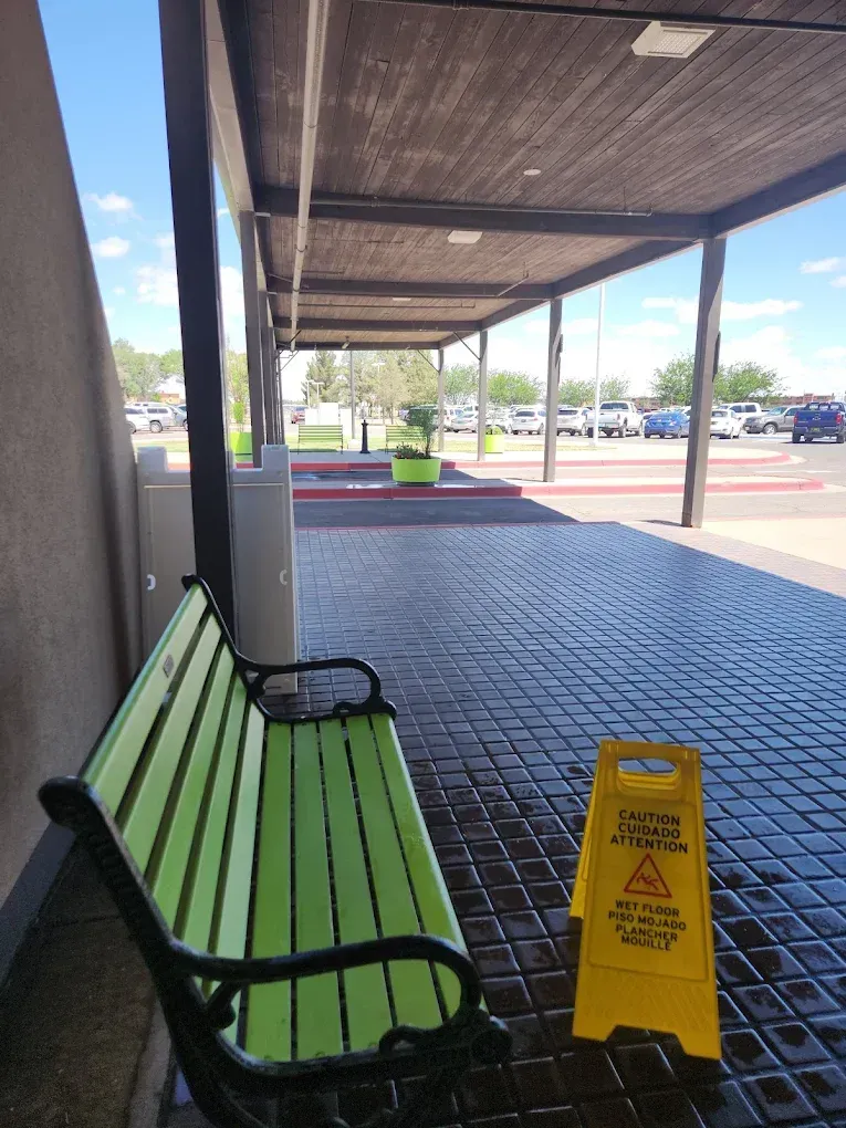Green bench beside a store wall with a yellow caution sign under a covered walkway outside.