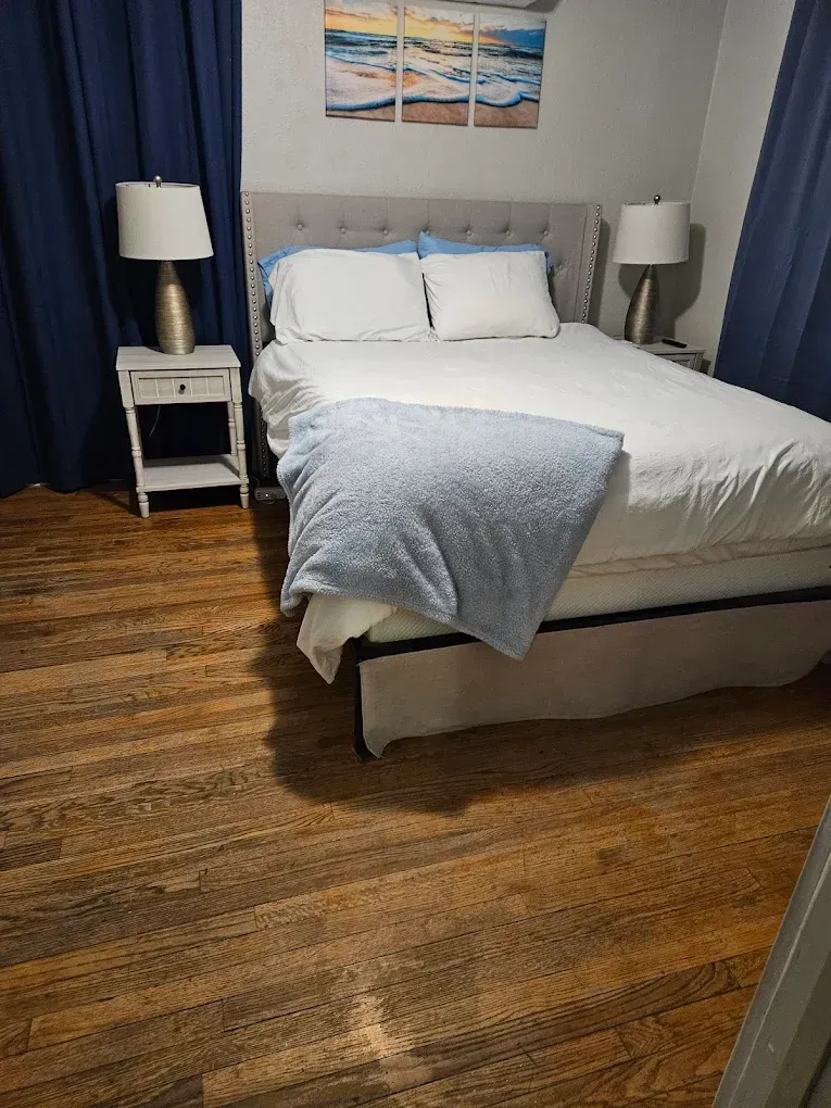 Bedroom with a made bed, gray headboard, bedside lamps, blue curtains, and wood flooring.