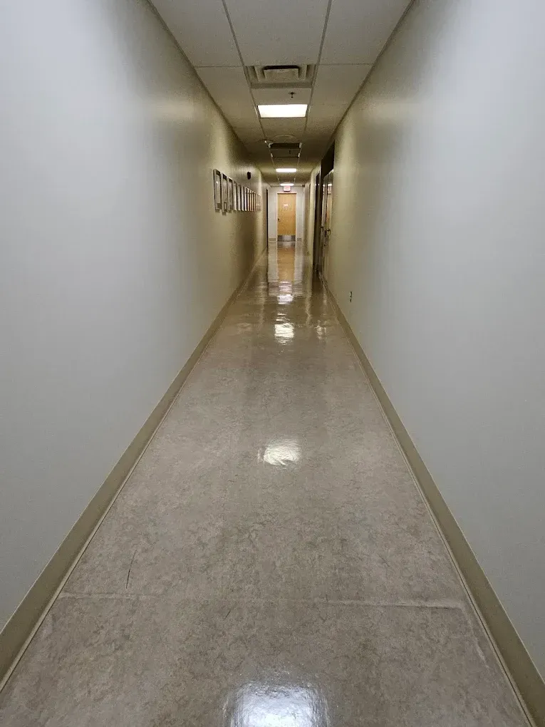 Empty indoor hallway with white walls, beige floor, and fluorescent ceiling lights, viewed toward a distant door