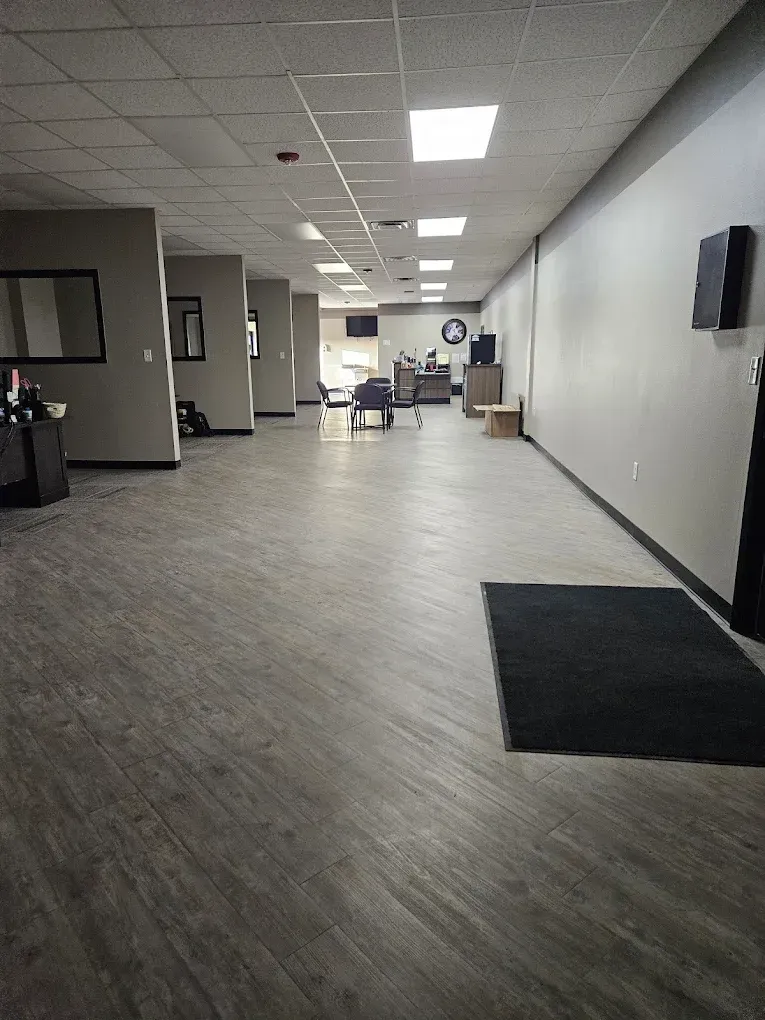 Office hallway with gray carpet, white walls, and ceiling lights, leading to a seating area at the far end