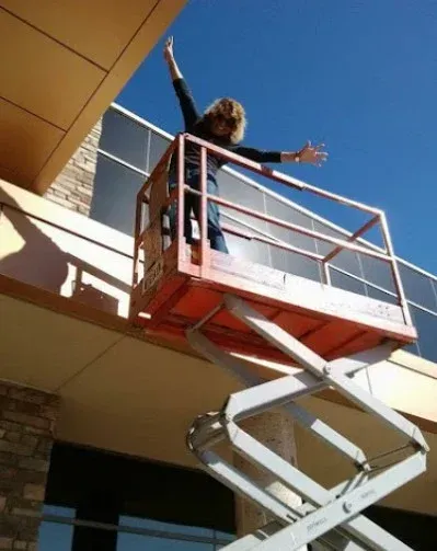 Person standing on a scissor lift with arms raised beside a building wall
