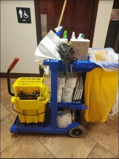 Janitorial cart with cleaning supplies, yellow mop bucket, and trash bags in a hallway