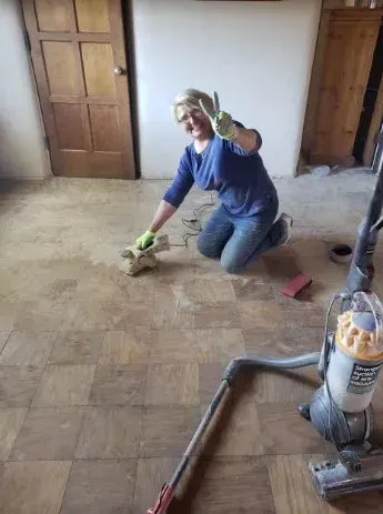 Person kneeling and cleaning a dusty floor with a cloth in an unfinished room, near a vacuum cleaner