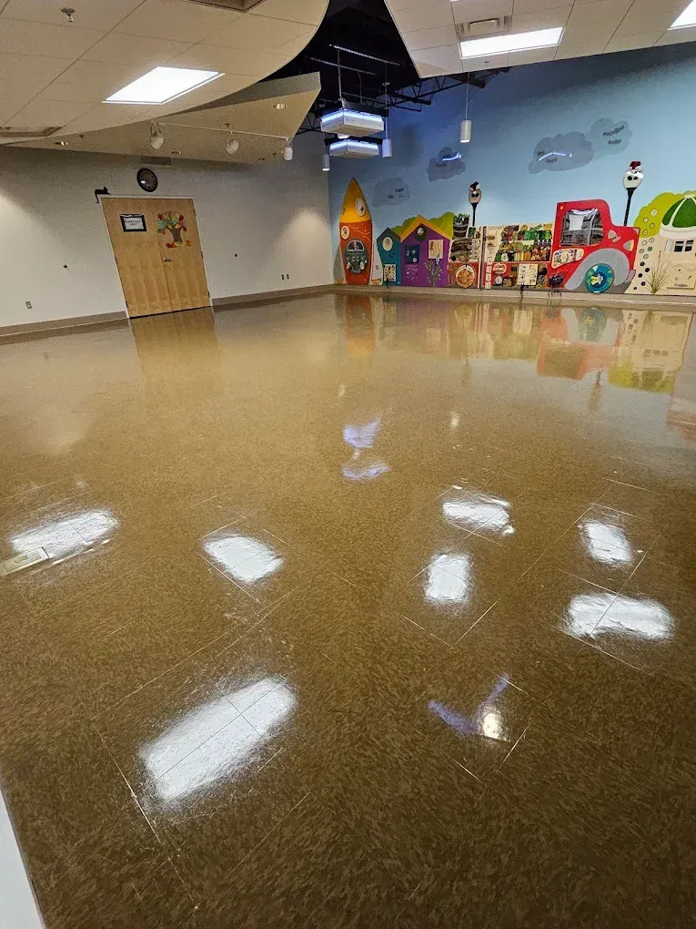 Empty community room with glossy floor, tan walls, and colorful children’s play equipment along the back wall