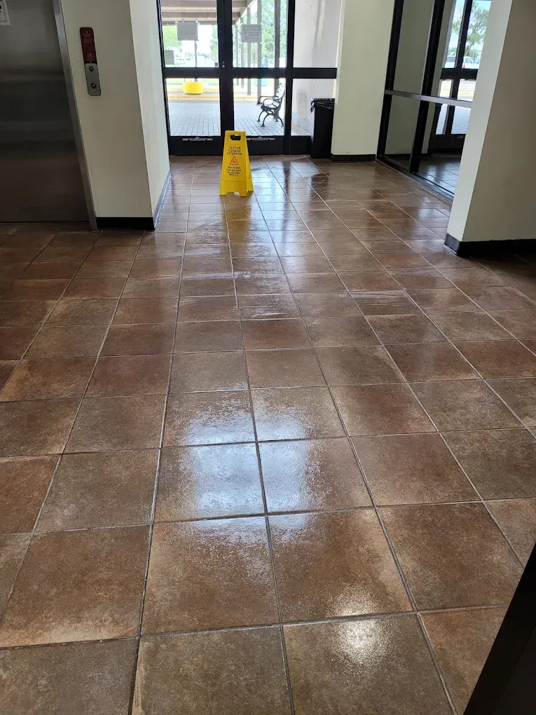 Indoor lobby with wet brown tile floor and a yellow caution sign near the glass doors