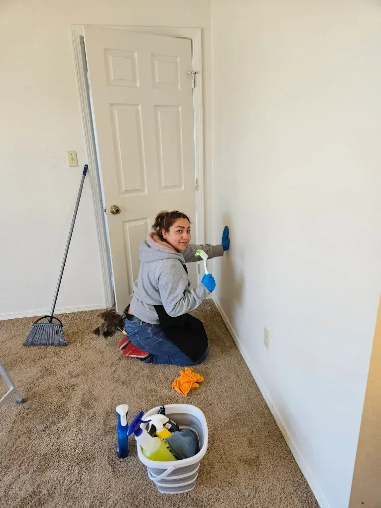 Person kneeling and cleaning a white wall with spray bottle, broom and bucket on carpeted floor.