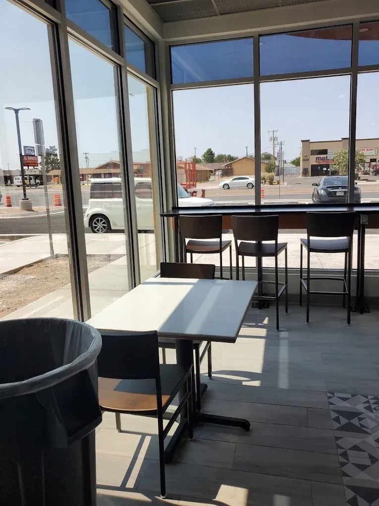 Empty café seating area with tables and stools by large windows, looking out to a sunny street.