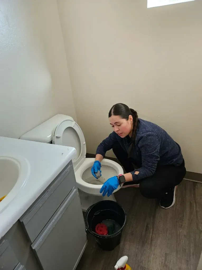 Person cleaning a toilet in a laundry room with a blue brush, black bucket, and washing machine nearby