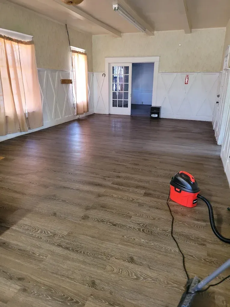 Empty room with wood flooring, beige walls, sheer curtains, and a red vacuum cleaner on the floor