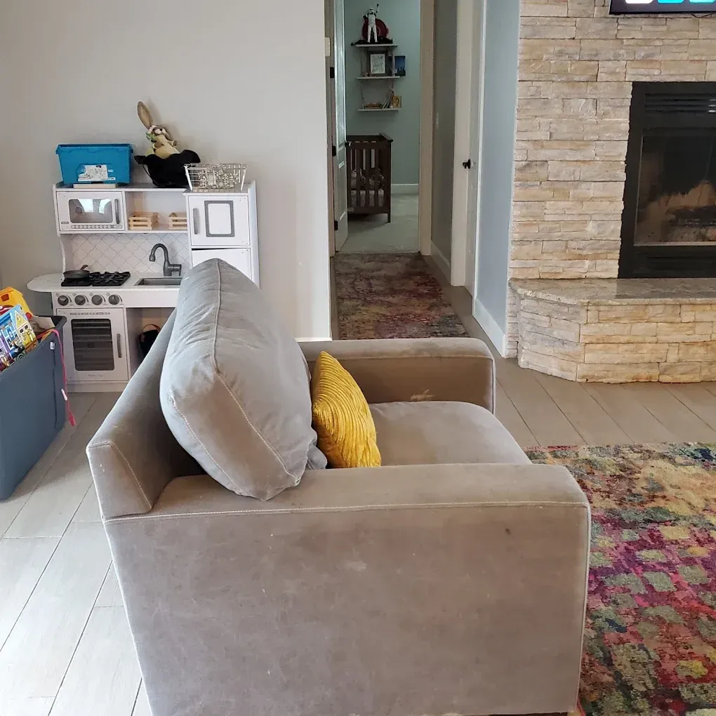 Modern living room with beige armchair, colorful rug, brick fireplace, and toy shelf in the background