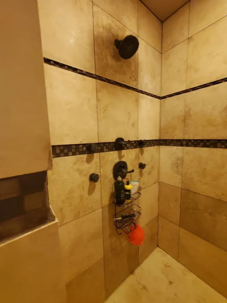 Bathroom shower stall with beige tile, a wall-mounted showerhead, and plumbing exposed at the center.
