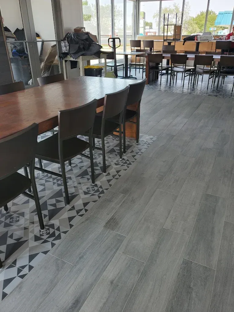 Modern cafeteria with long tables, black chairs, and patterned tile flooring near large windows.