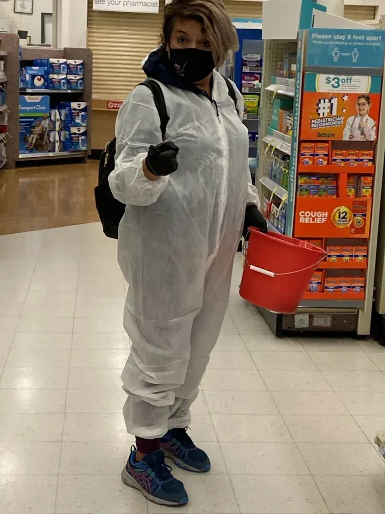 Person in white protective coveralls, black mask and gloves, holding a bucket in a store aisle.