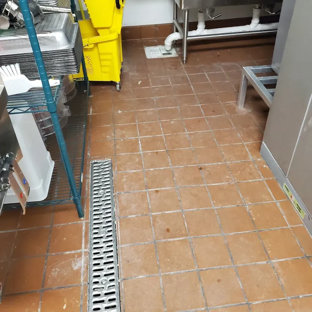 Tiled kitchen floor with a metal drain grate, cleaning cart, and storage shelves along the wall