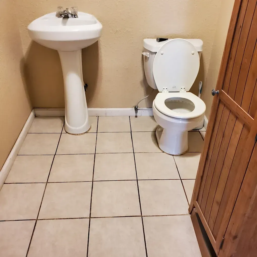 Small restroom with white pedestal sink, toilet, tan walls, tiled floor, and wooden stall door.
