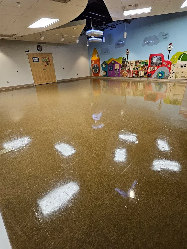 Empty community room with glossy floor, tan walls, and colorful children’s play equipment along the back wall