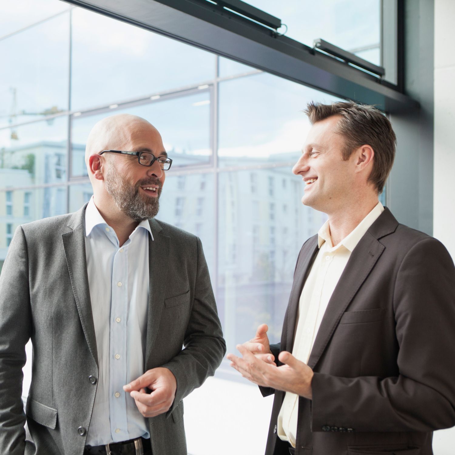Two men in suits are talking in front of a window