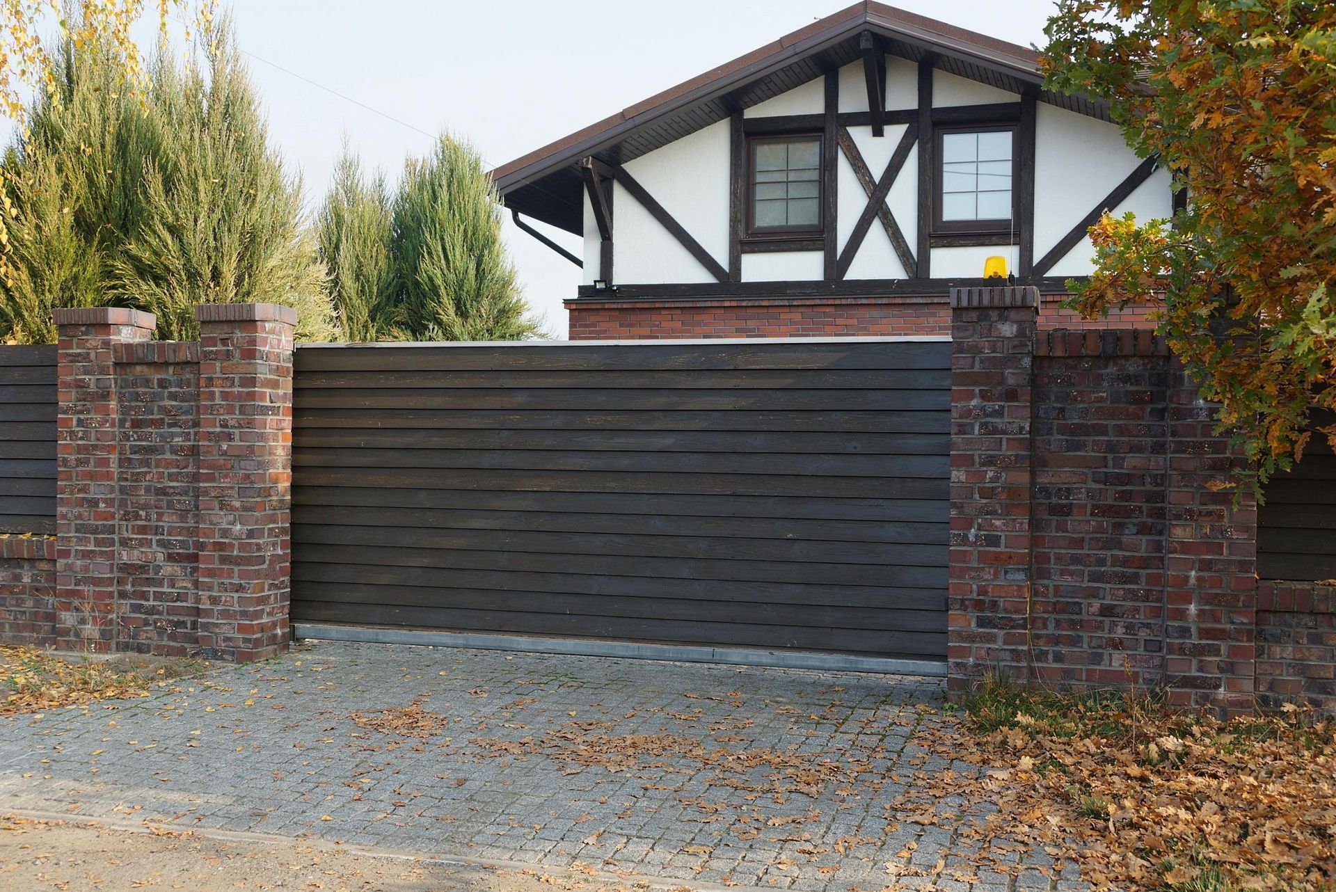Dark wooden gate set between brick columns, in front of a house with a white facade and dark trim.