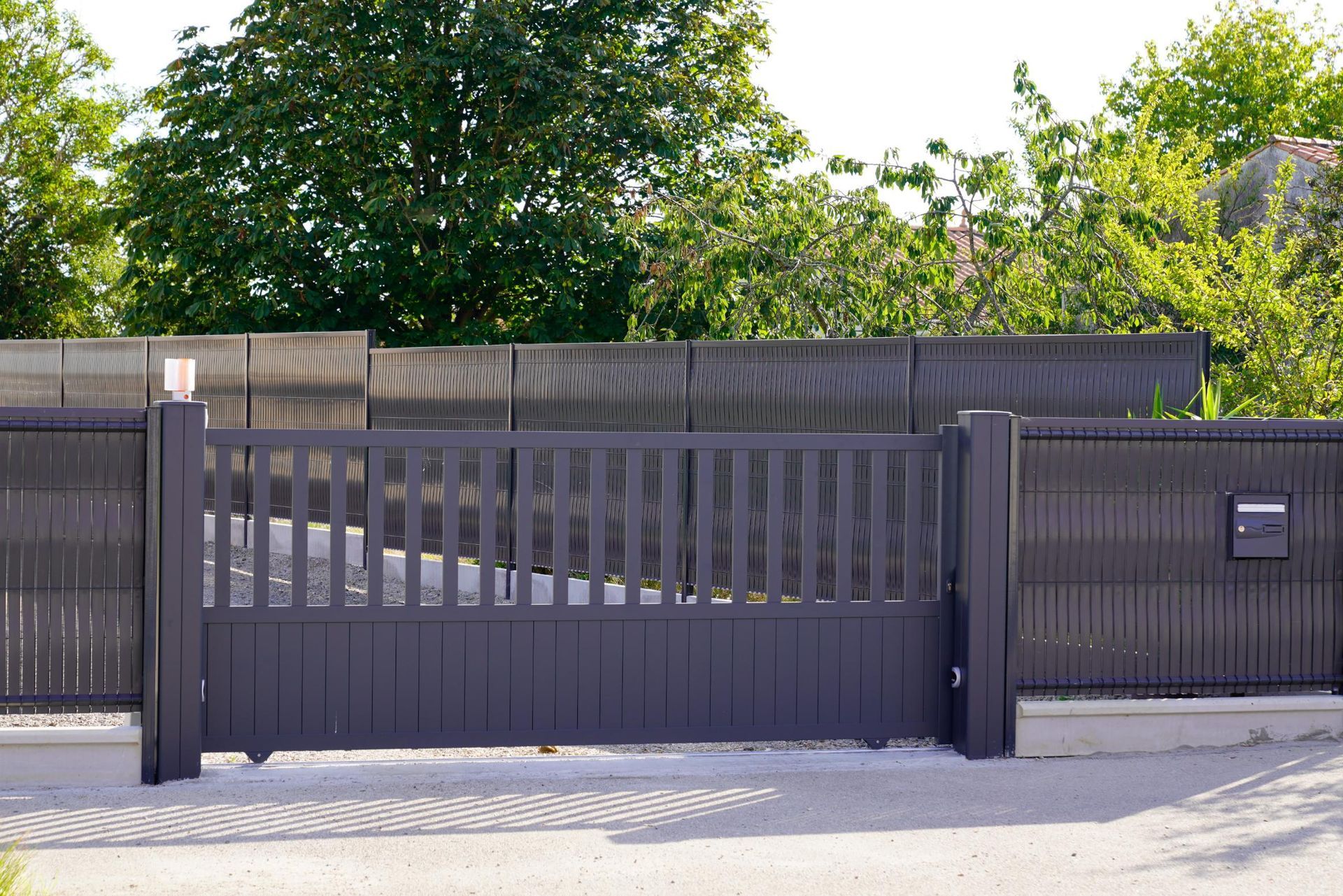 Dark gray metal gate, fence, and mailbox in front of a house. Trees in background.