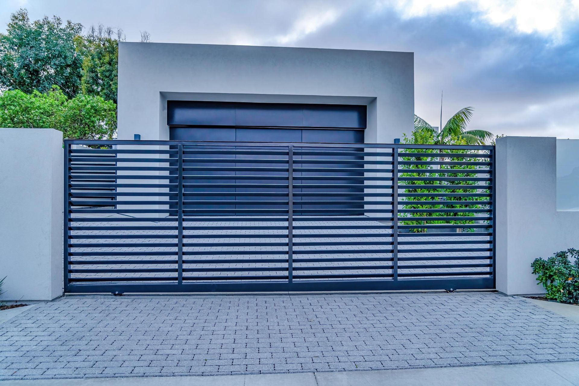 Modern gray gate in front of a white building with a dark garage door, cobblestone driveway.