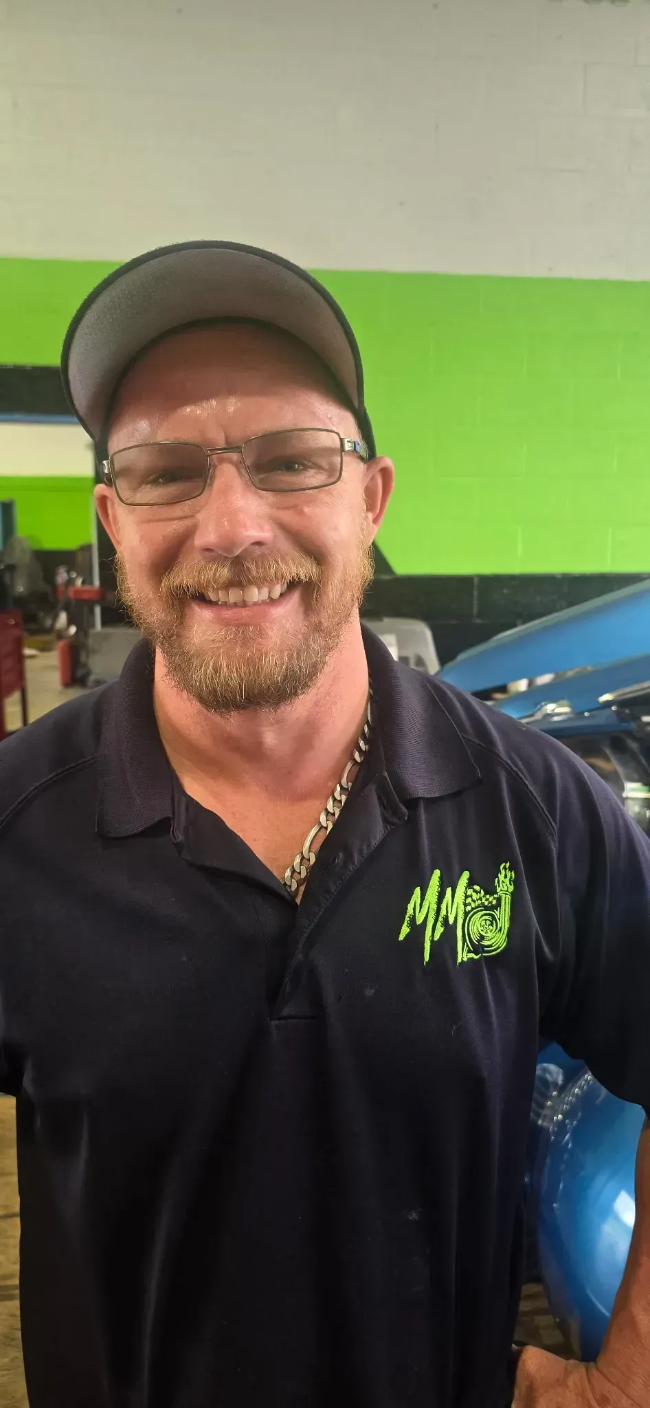 Smiling man in cap and glasses standing beside a blue car in a garage