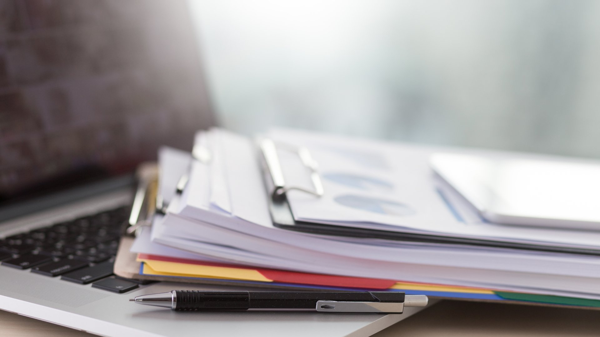Laptop with stacked notebooks, folders, and a pen on a desk in soft light