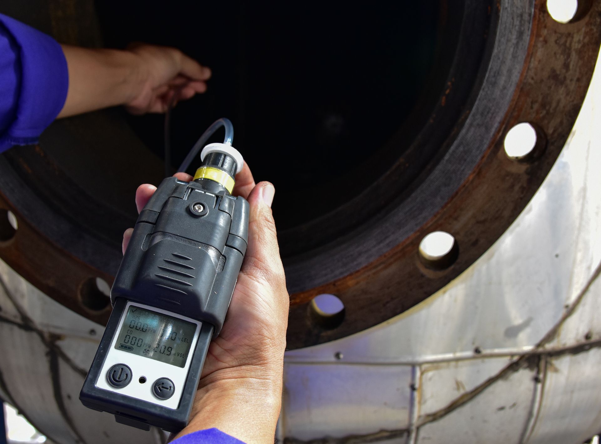 Hand holding a handheld portable gas detector inside a large metal pipe opening.