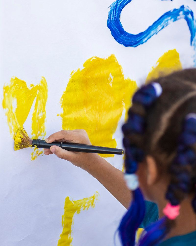 Child painting yellow shapes on a white wall with a brush, with blue paint visible above.
