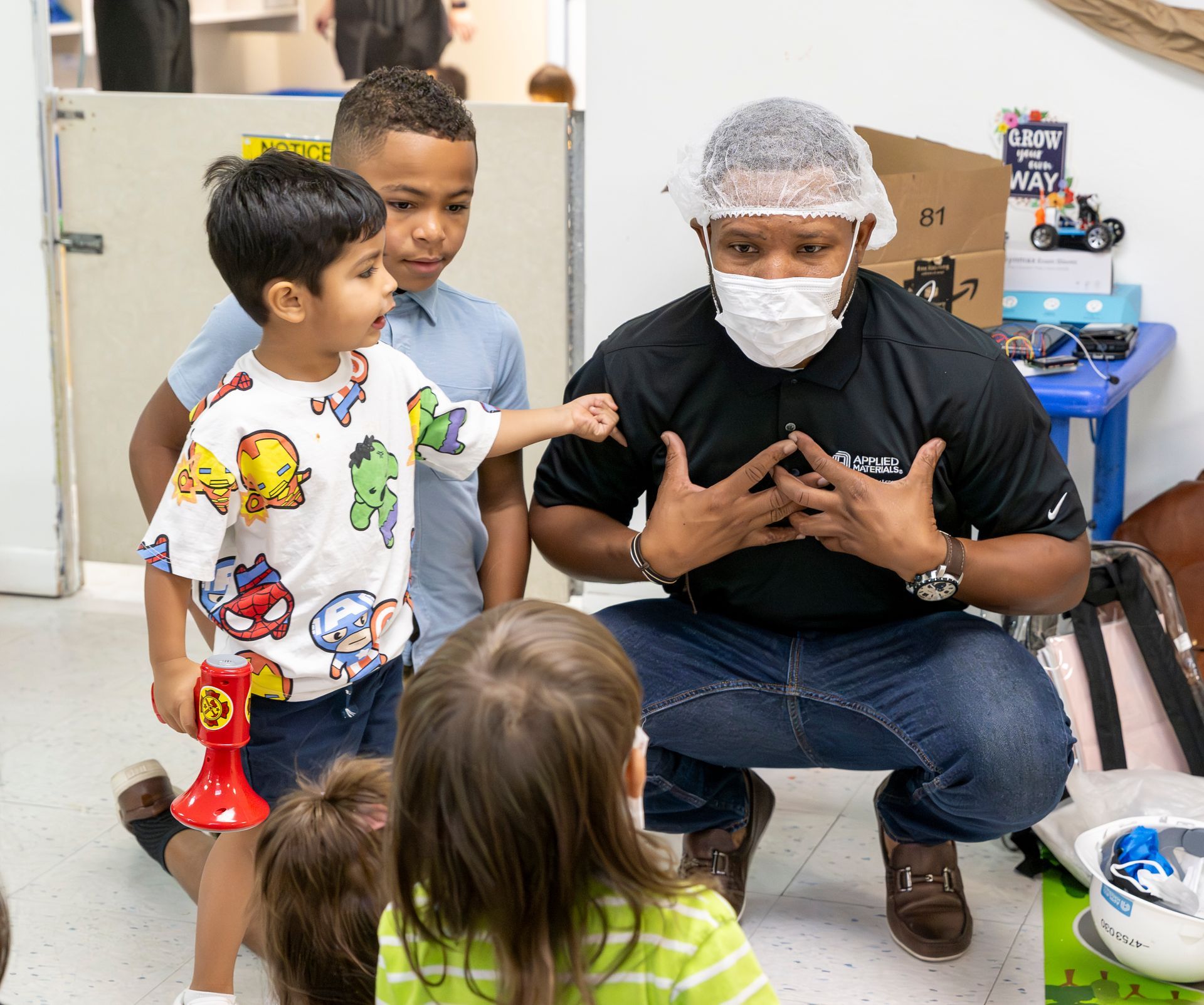 Adult wearing a mask and hair net kneels with children in a classroom, engaging at eye level.