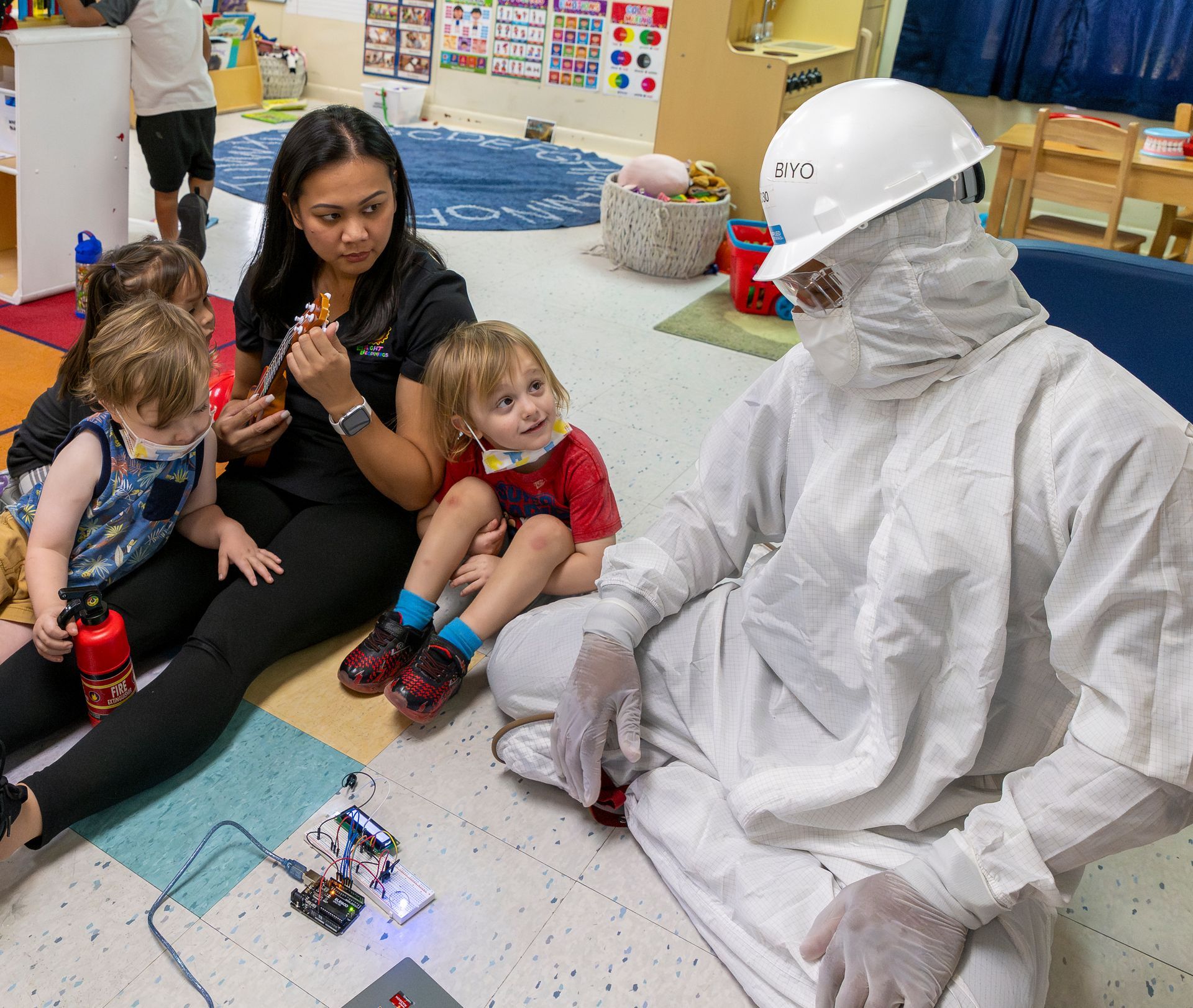 Children sit on the floor with an adult in a white protective suit and hard hat in a classroom.