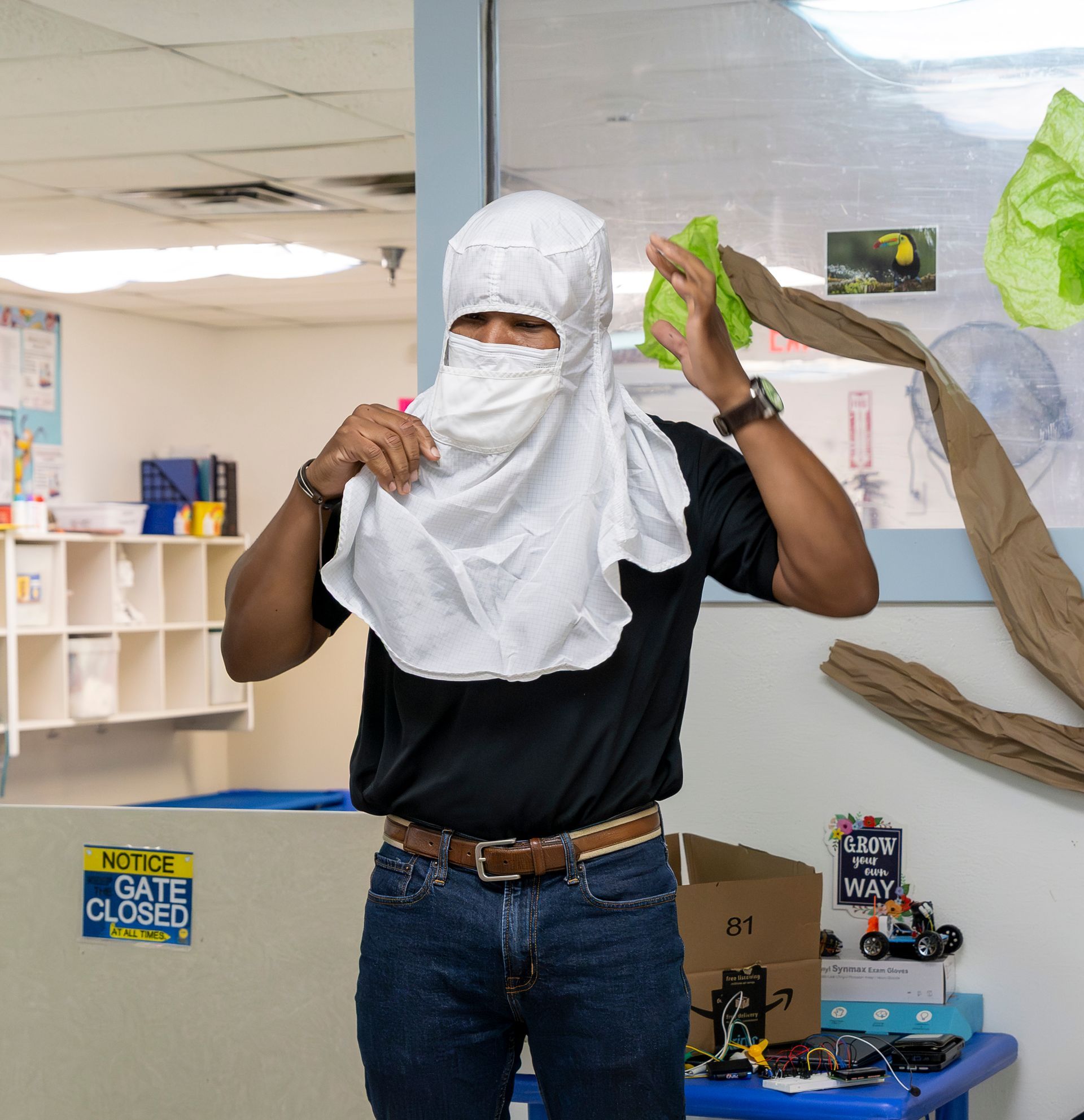 Person in a black shirt adjusts a white cloth head covering in a classroom or play area.