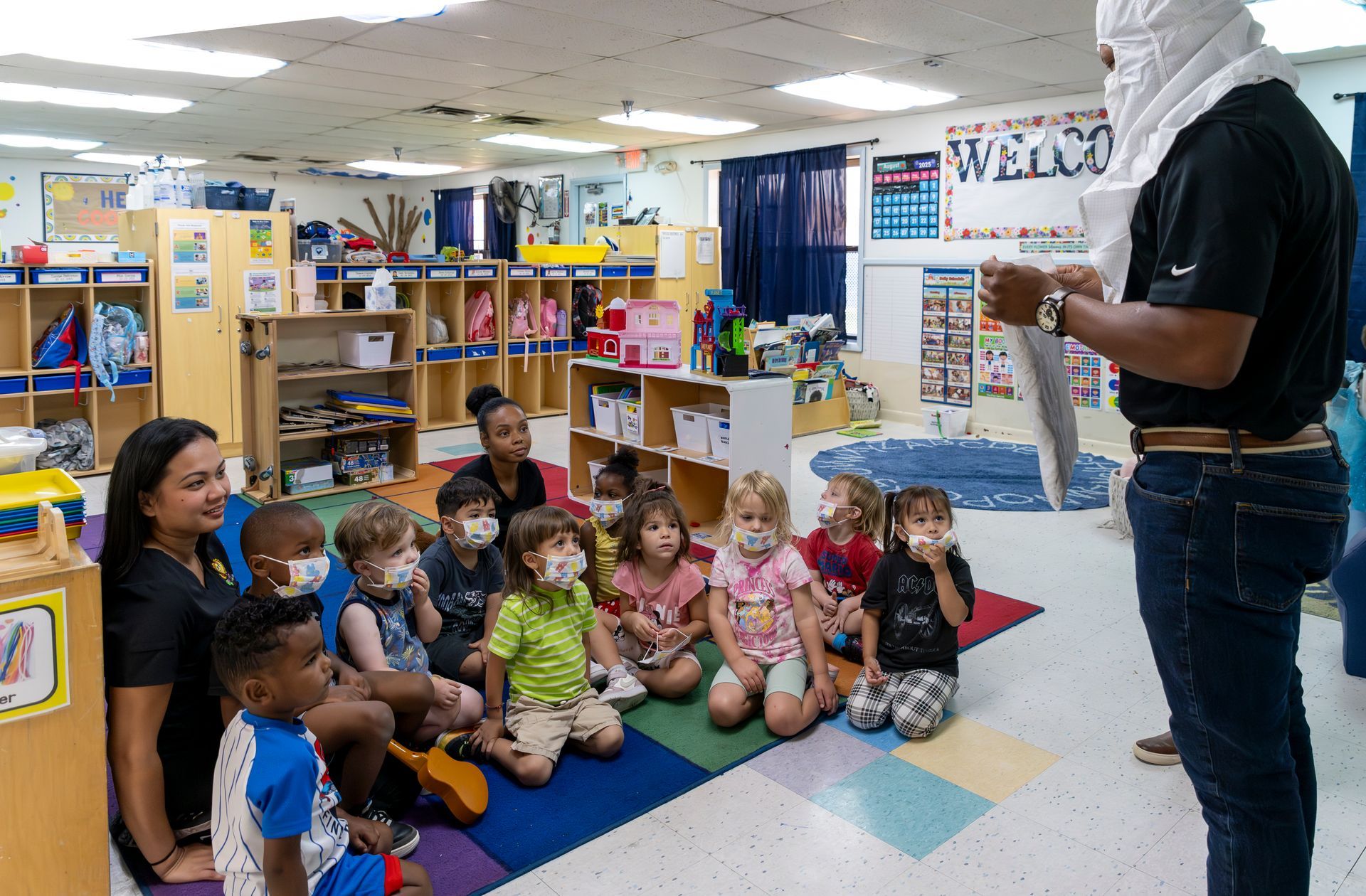 Teacher stands before seated children in a colorful classroom, holding papers during a lesson.