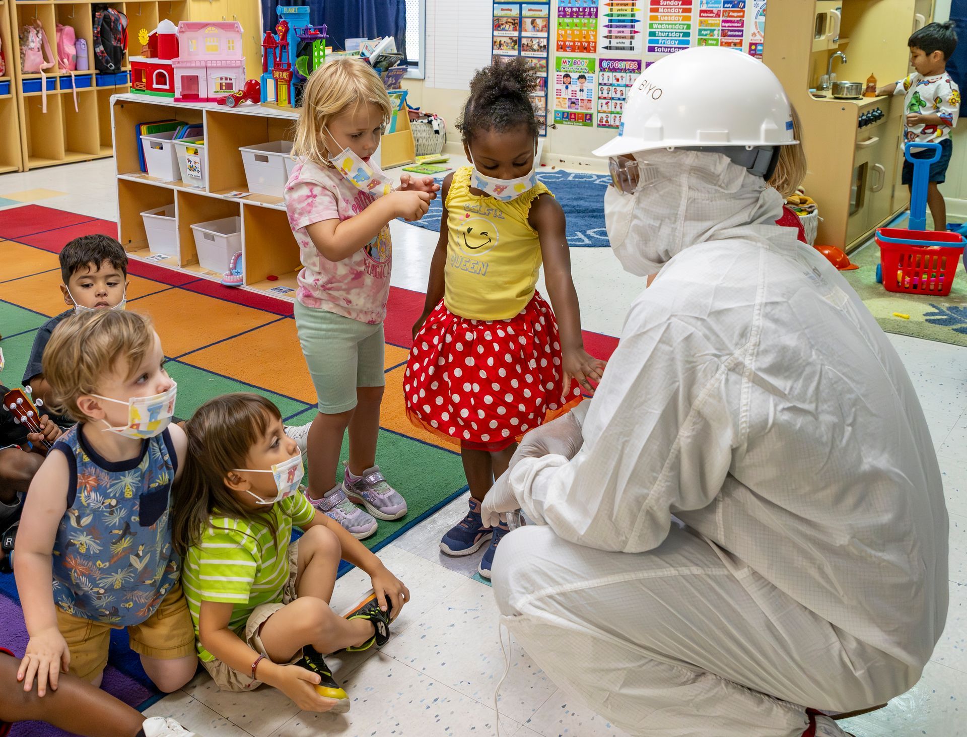 Children in a classroom watching a worker in white protective gear and face shield.