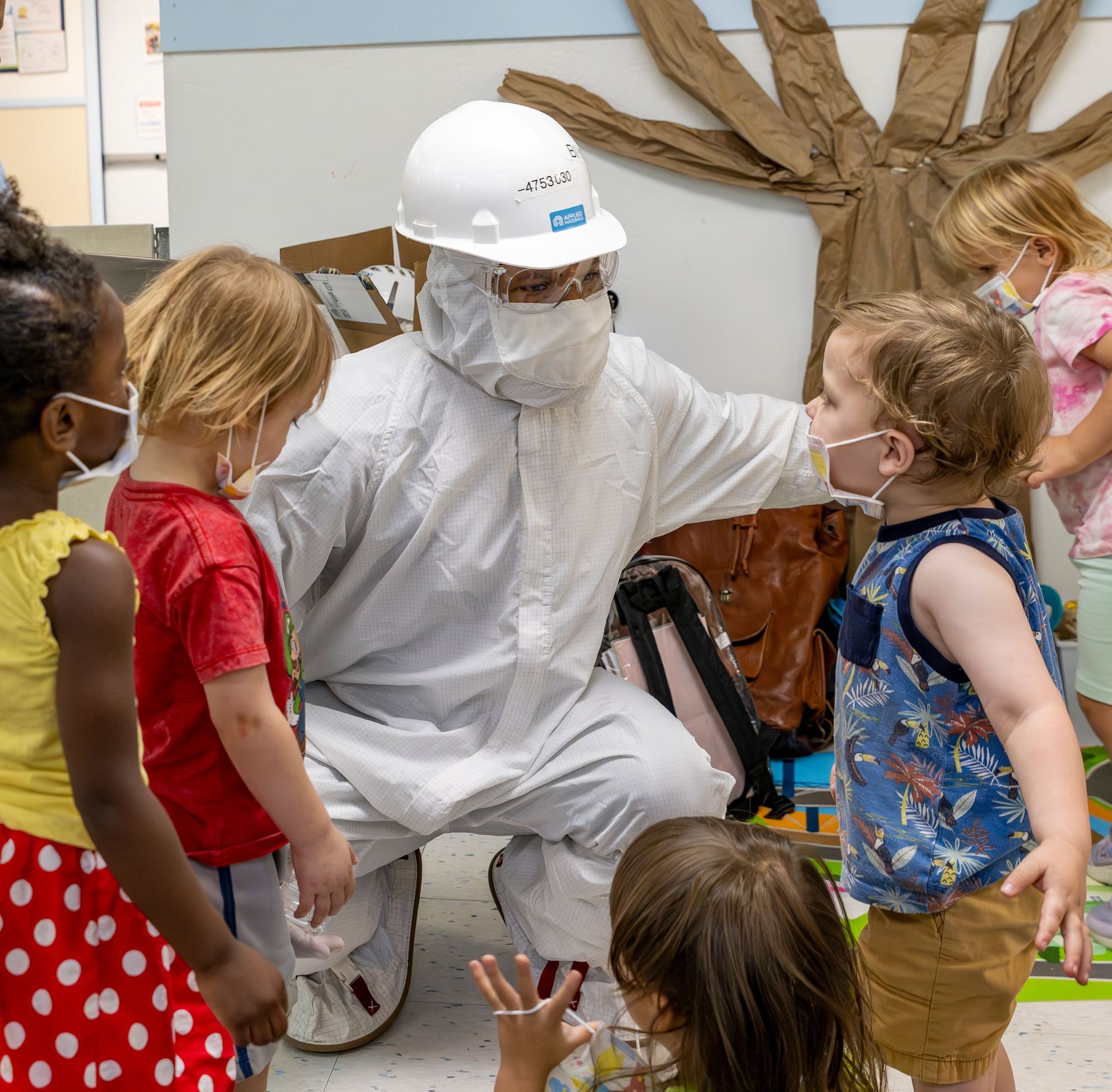 Worker in white protective suit kneels with children in a classroom, showing them a brown object.