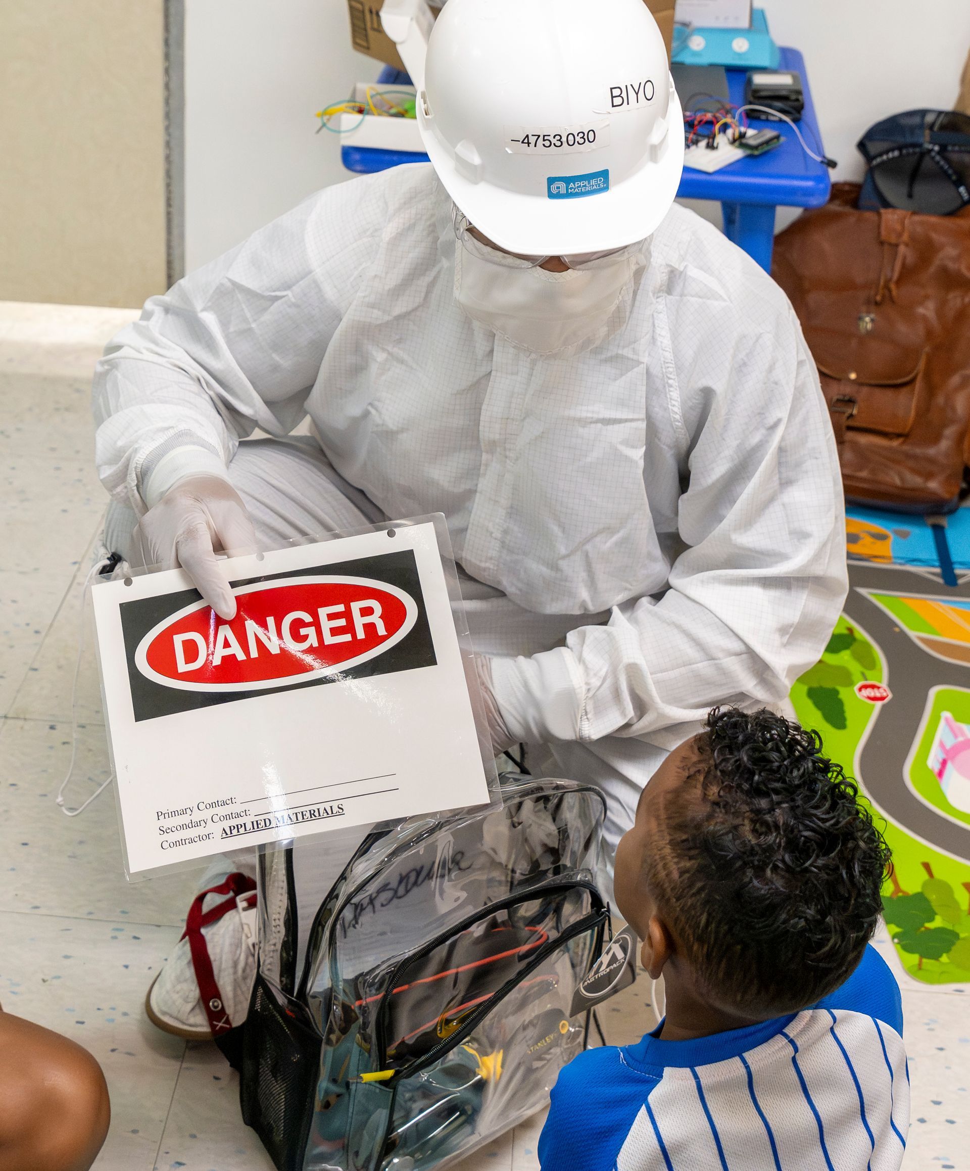 Worker in white protective gear holding a DANGER sign near exposed wiring while a child watches.