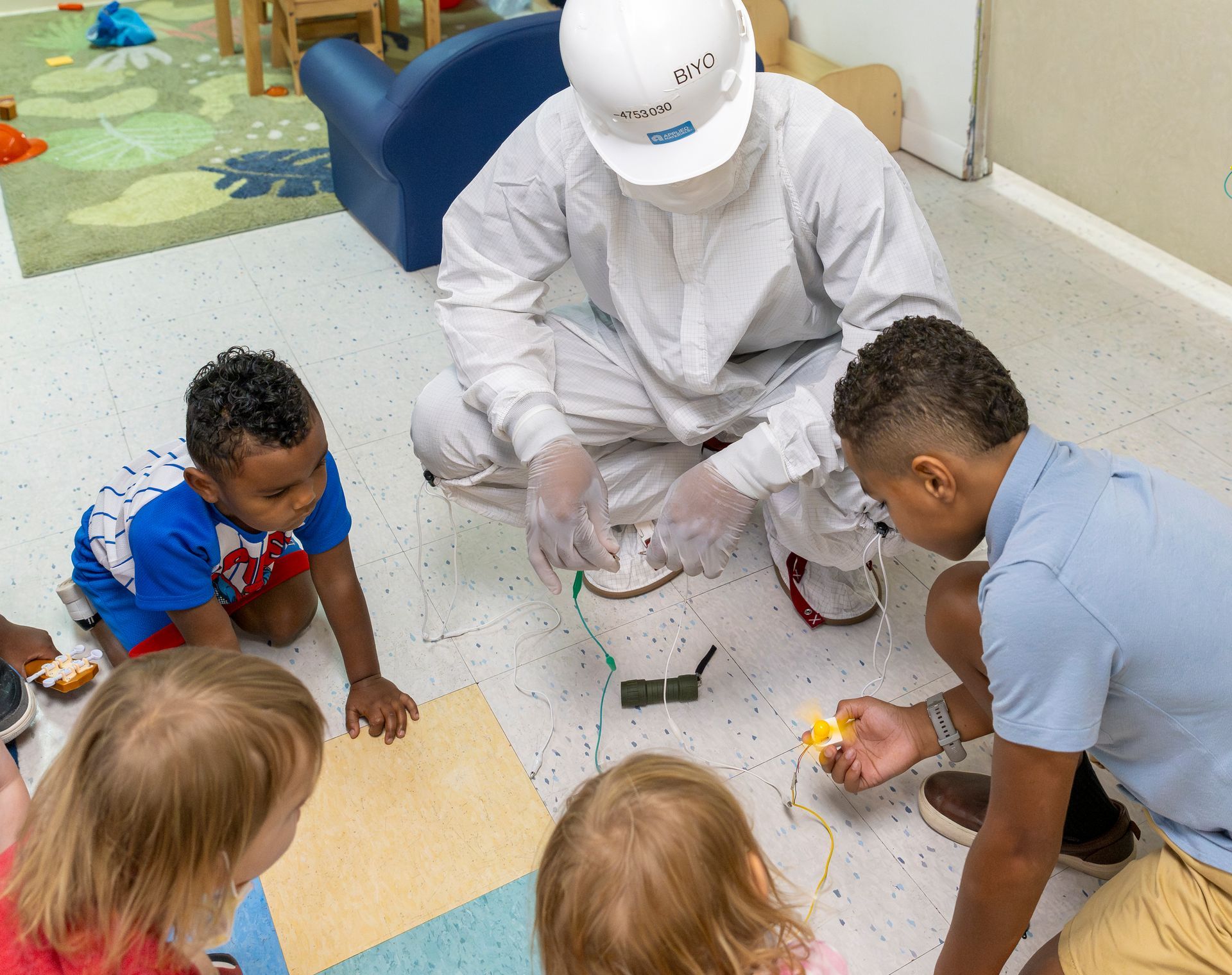 Children and an adult in protective gear build a small device on the floor in a classroom.