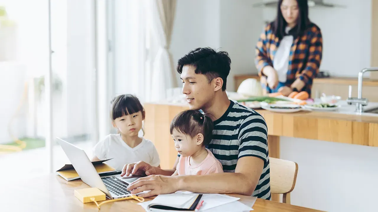 Family working together at a kitchen table with a laptop and papers, while another person cooks in the background