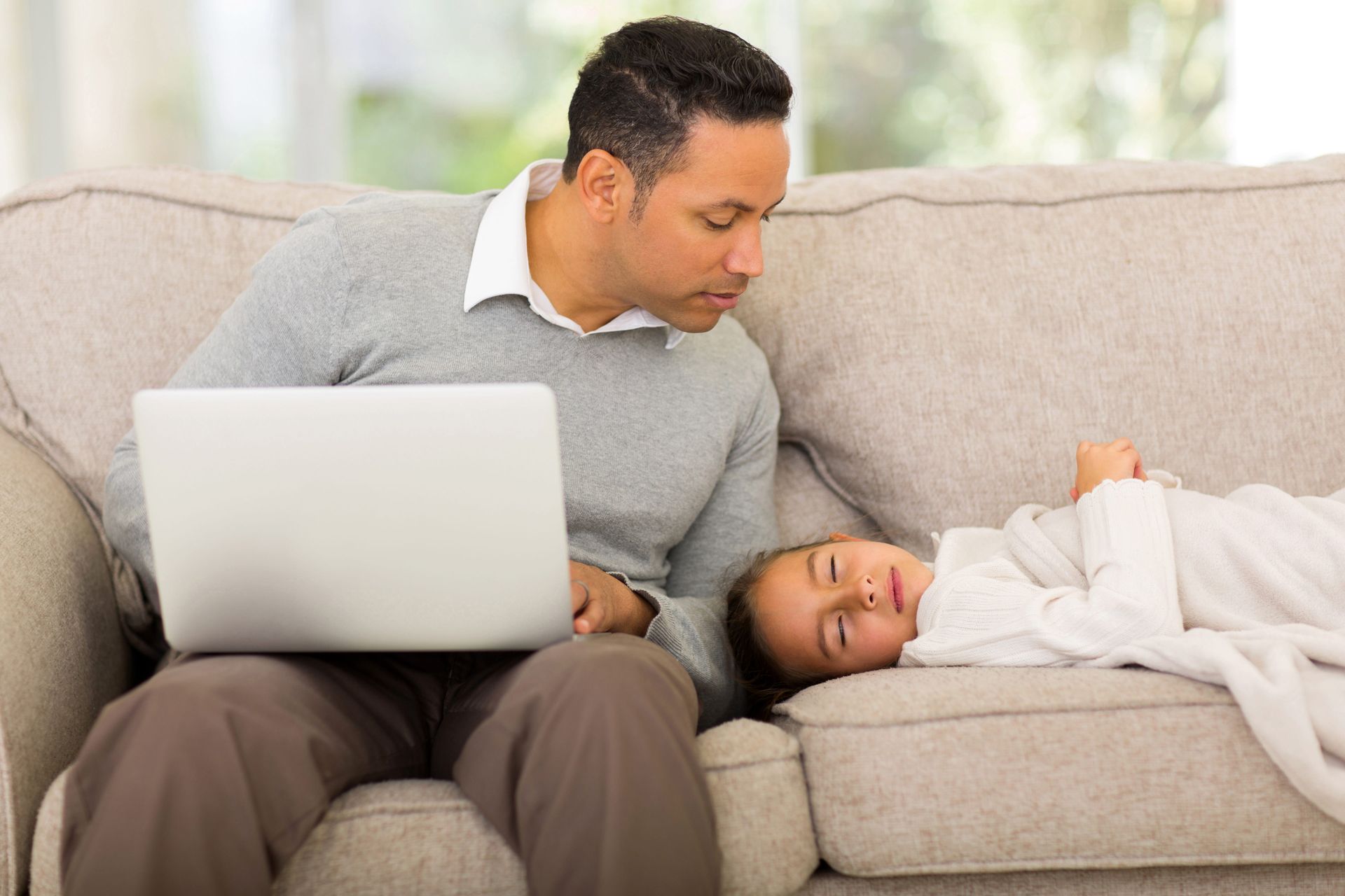 Man on a couch with a laptop beside a child resting under a white blanket