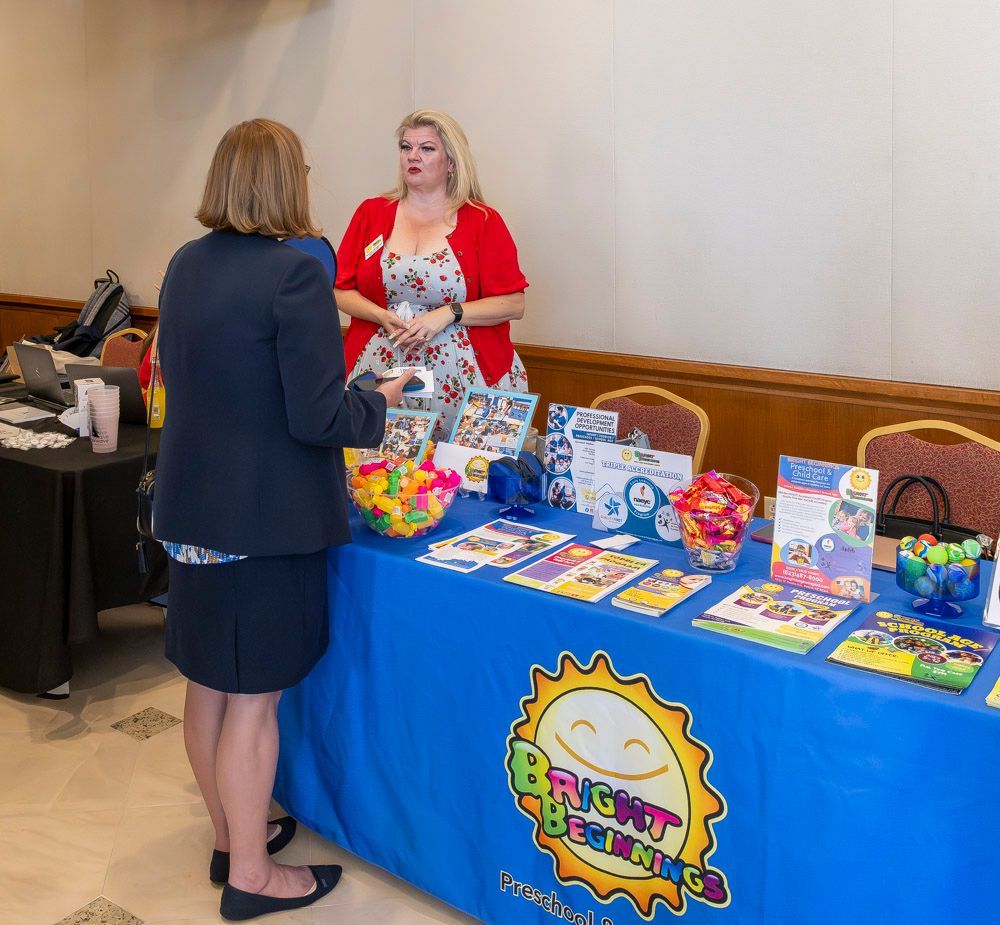 Two women at a bright birthday-themed vendor table with colorful gift items and cards.