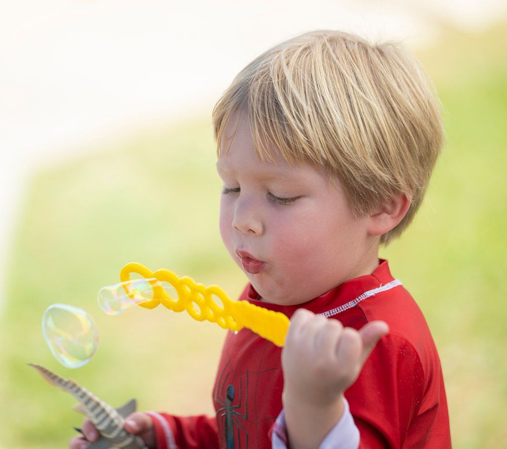 Child blowing bubbles outdoors, holding a bubble wand and tray, wearing a red shirt.