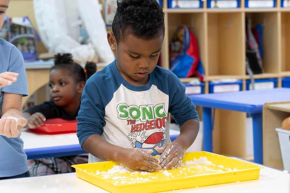 Child in a classroom kneads dough on a yellow tray while another child watches in the background.
