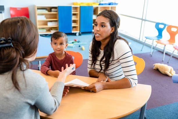 Three people talking at a table in a colorful classroom, with a child between two adults.