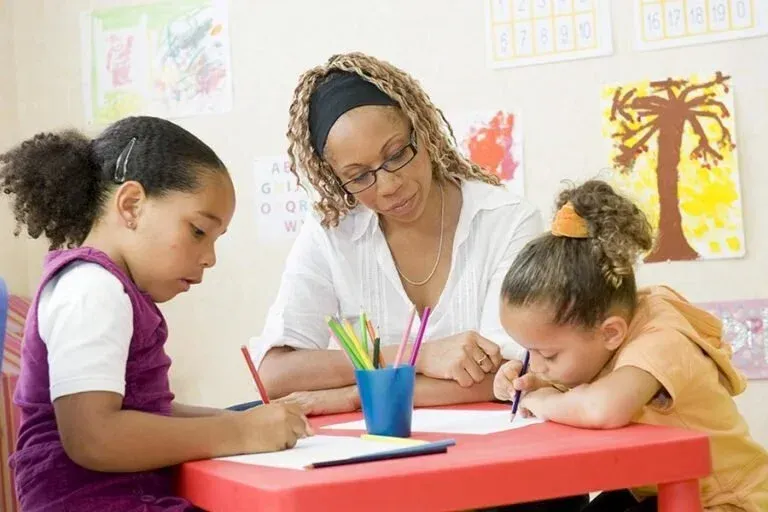 Teacher helping two children draw at a red table with colored pencils in a classroom