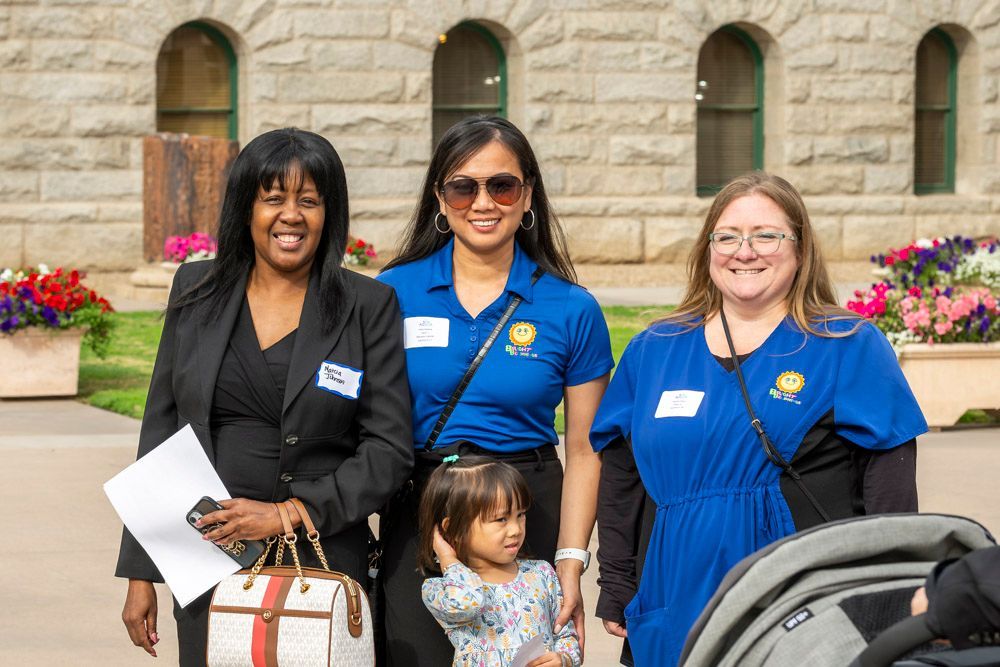 Three women and a child stand outdoors in front of a stone building with flower planters.