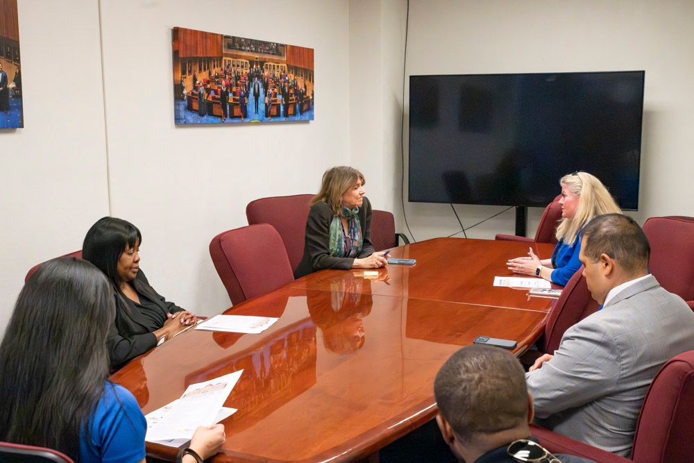 People seated around a conference table in a meeting room, talking with a wall-mounted TV nearby