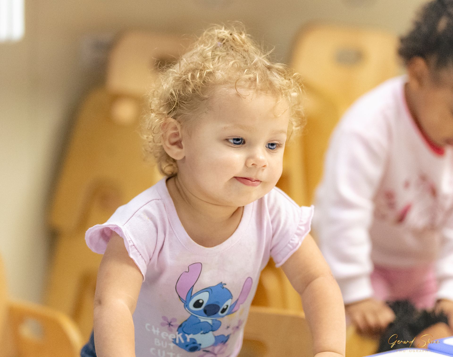 Toddler in a pink shirt with a cartoon graphic, leaning forward in a bright classroom.