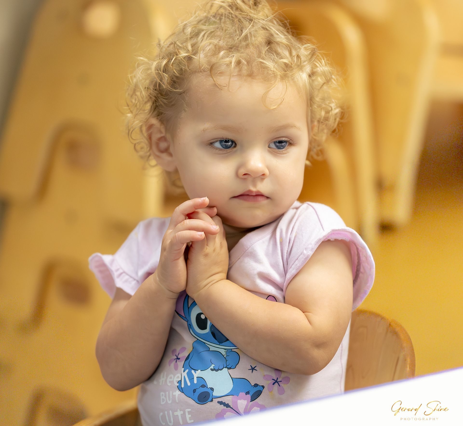 Young child with curly blonde hair in a pink shirt, clasping hands at a table, looking to the side