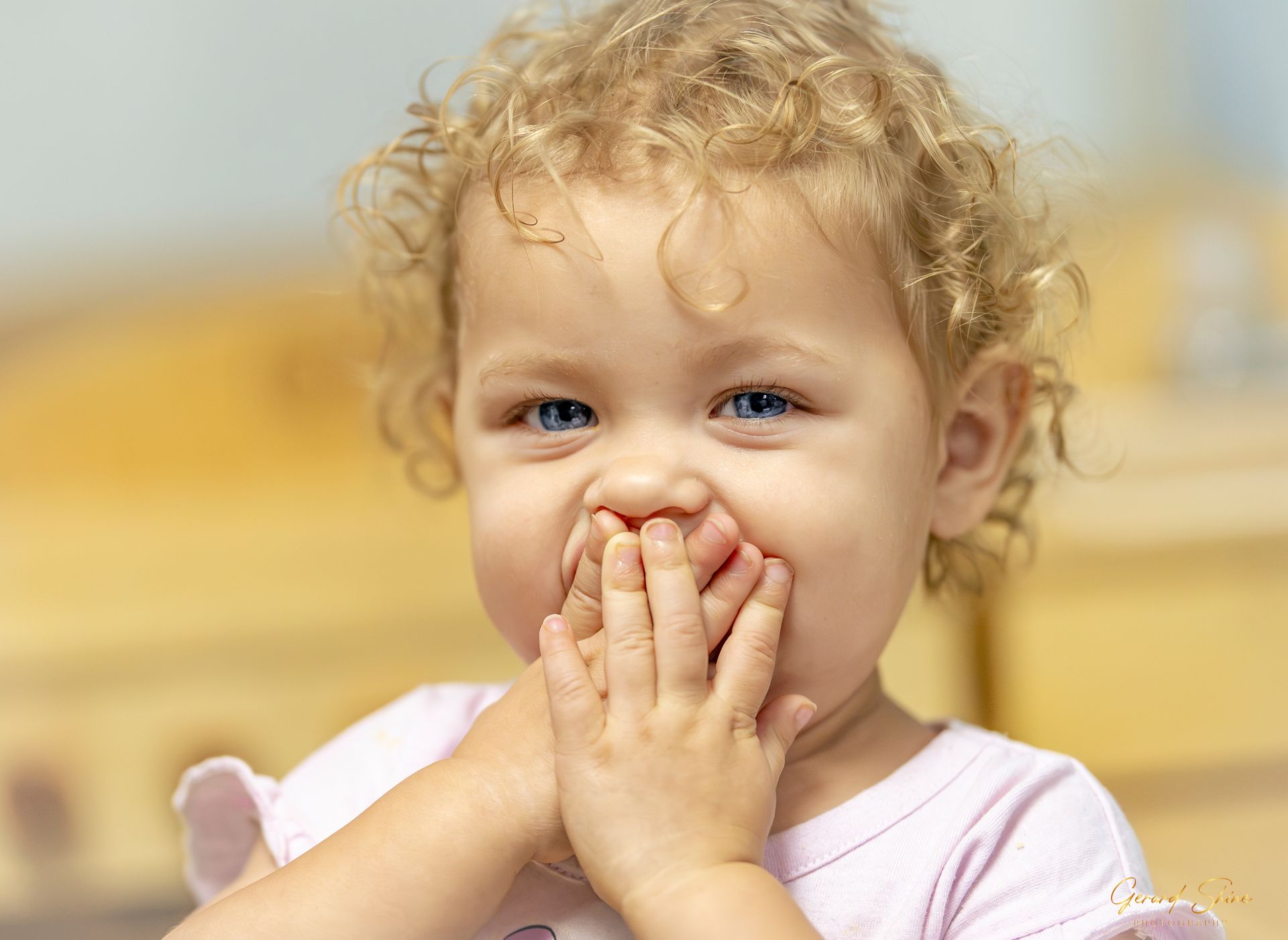 Smiling curly-haired child covering mouth with hands, in a light pink shirt on a blurred indoor background