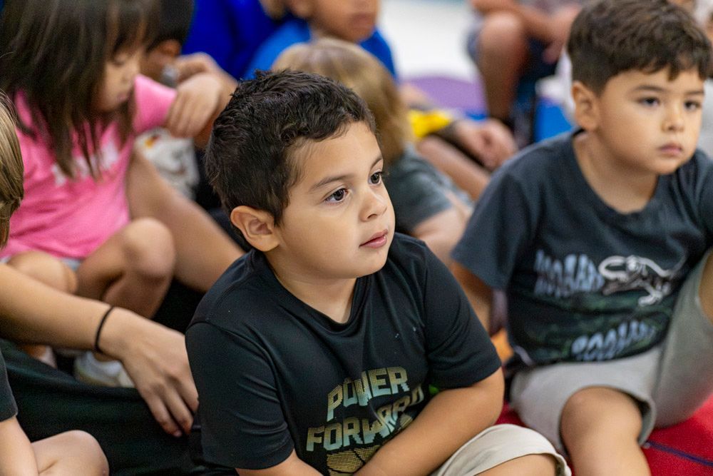 Children sitting cross-legged on a classroom floor, listening attentively during an activity