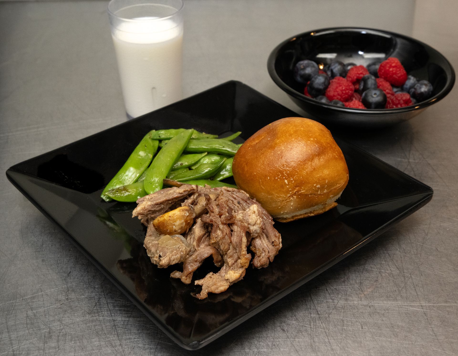Plate with steak, green peppers, and a bread roll, with a glass of milk and berries in the background